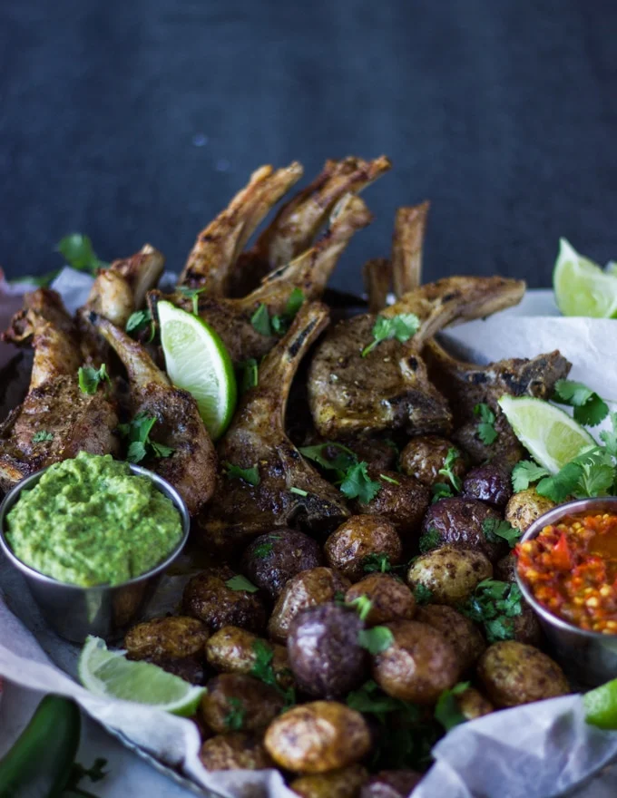 A plate showing crispy air fryer potatoes next to air fryer lamb chops and the poblano sauce in a bowl, sprinkled with cilantro and lime slices
