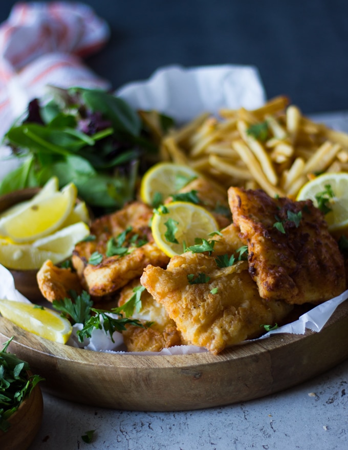 A finished plate of fish fry and fries on a wooden board surrounded by lemon slices and sprinkled with parsley