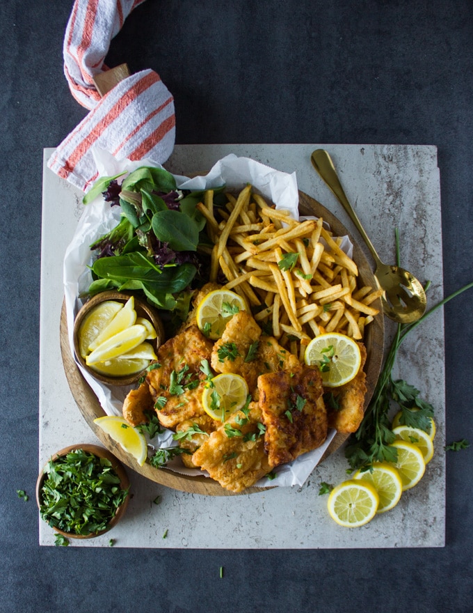 A huge platter with fish fry, lemon wedges, french fries and salad on a wooden board over a marble surrounded by lemon slices and a bowl of parsley