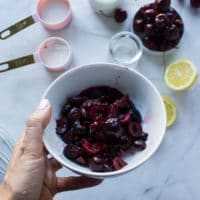 A hand holding a bowl of frozen cherries to make cherry pie filling with the remaining ingredients in a white marble below.