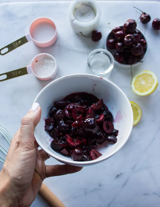A hand holding a bowl of frozen cherries to make cherry pie filling with the remaining ingredients in a white marble below.