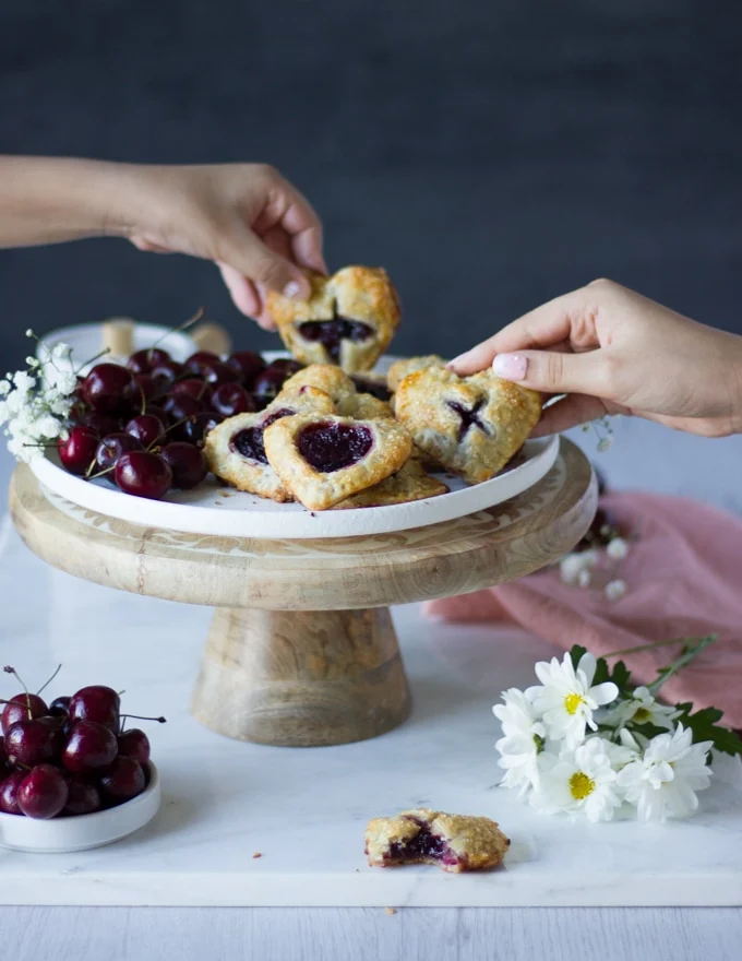 two kids hands grabbing the cherry pies from a serving plate on a white marble surrounded by white flowers and a pink tea towel