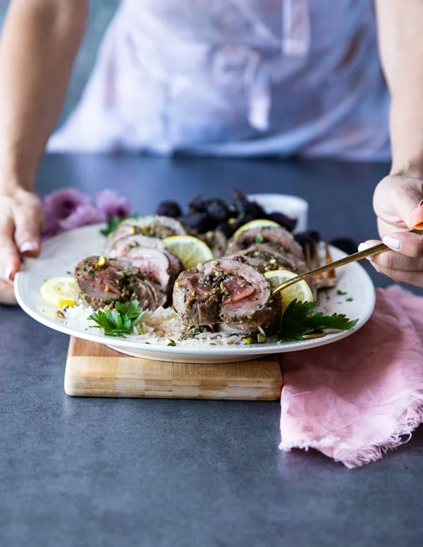 Two hands holding a plate of roast boneless leg of lamb over rice, sliced thinly and one hand serving the lamb using a spoon