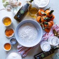 A list of ingredients for the olive oil cake showing a bowl of flour, another bowl of sugar, a cup of olive oil, some clementine or orange zest and juice, a bowl of yogurt and a bowl of eggs, also jars of baking powder and baking soda