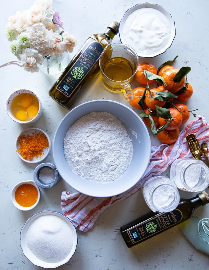 A list of ingredients for the olive oil cake showing a bowl of flour, another bowl of sugar, a cup of olive oil, some clementine or orange zest and juice, a bowl of yogurt and a bowl of eggs, also jars of baking powder and baking soda