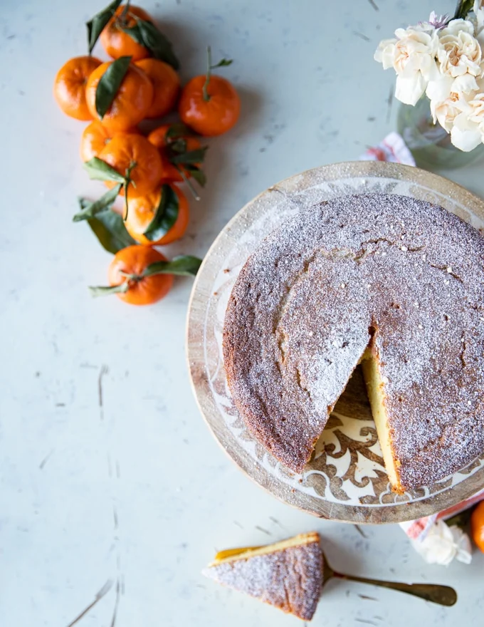 Top view of an orange olive oil cake with some clementine oranges on the side and some powdered sugar dusted over the cake