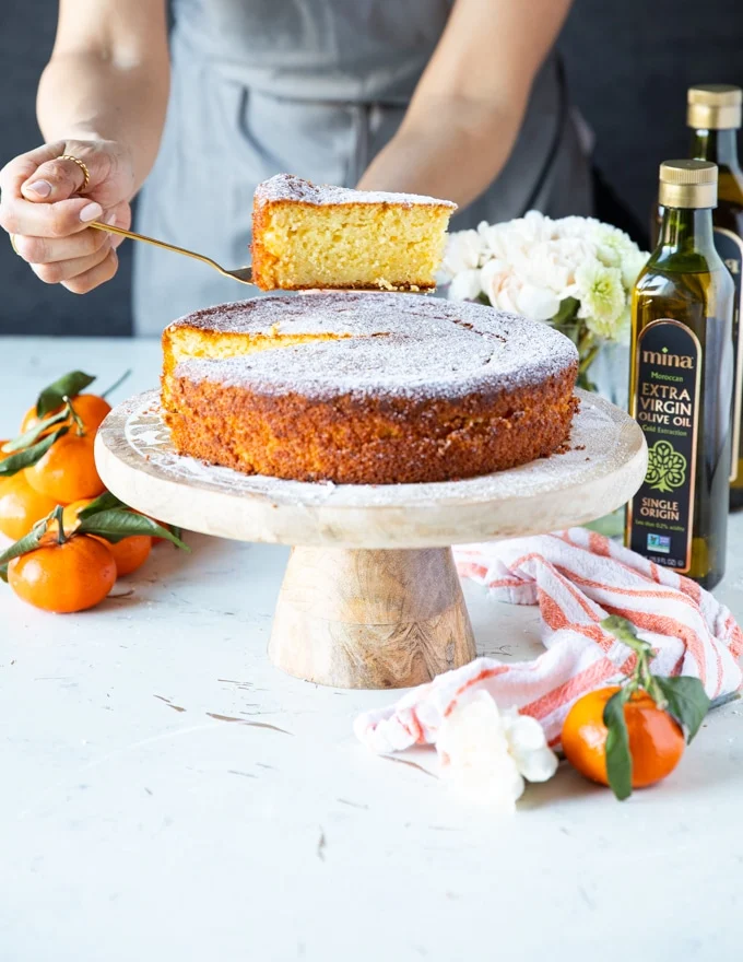 A hand holding a slice of olive oil cake over the serving olate with some clementine oranges and a tea towel around it
