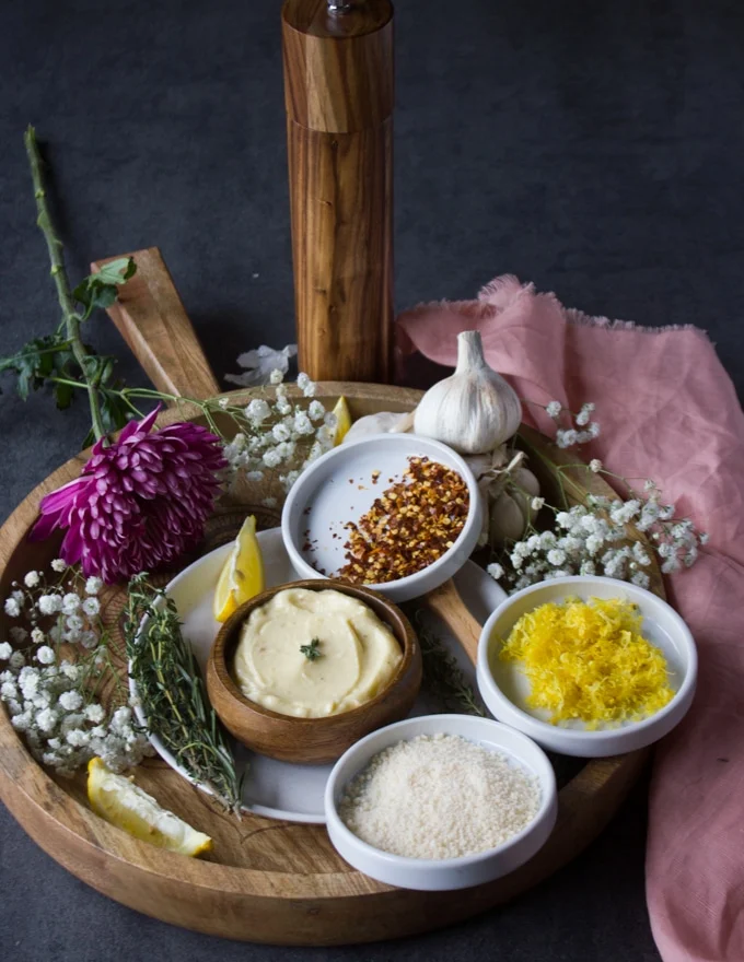The garlic butter sauce on a tray surrounded with small bowls of flavoring such as chilli flakes, lemon zest, parmesan cheese, herbs and lemon wedges