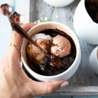 A hand holding a brownie in a mug loaded with scoops of chocolate and vanila ice cream and caramel sauce