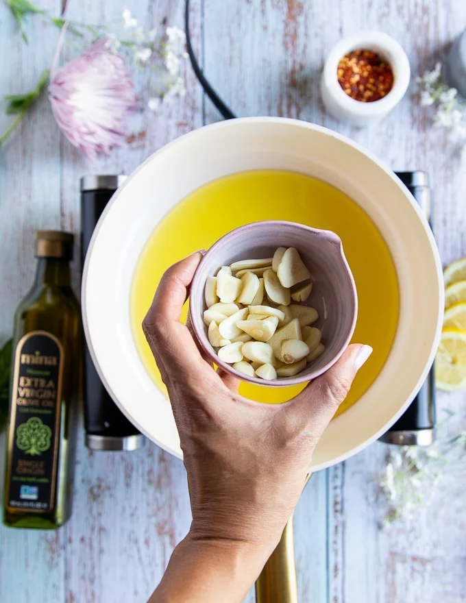 A hand holding the sliced garlic over a skillet of olive oil