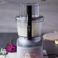 A hand drizzling oil in a thin stream through the feed of a food processor to make toum recipe