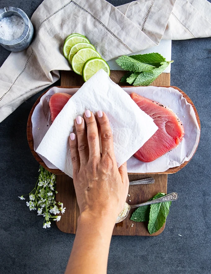 A hand holding a kitchen towel patting down the tuna steaks dry before seasoning