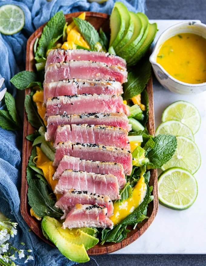 sliced tuna steak recipe on a bed of mint and greens surrounded by lime slices a tew towel and two salad spoons on a white marble