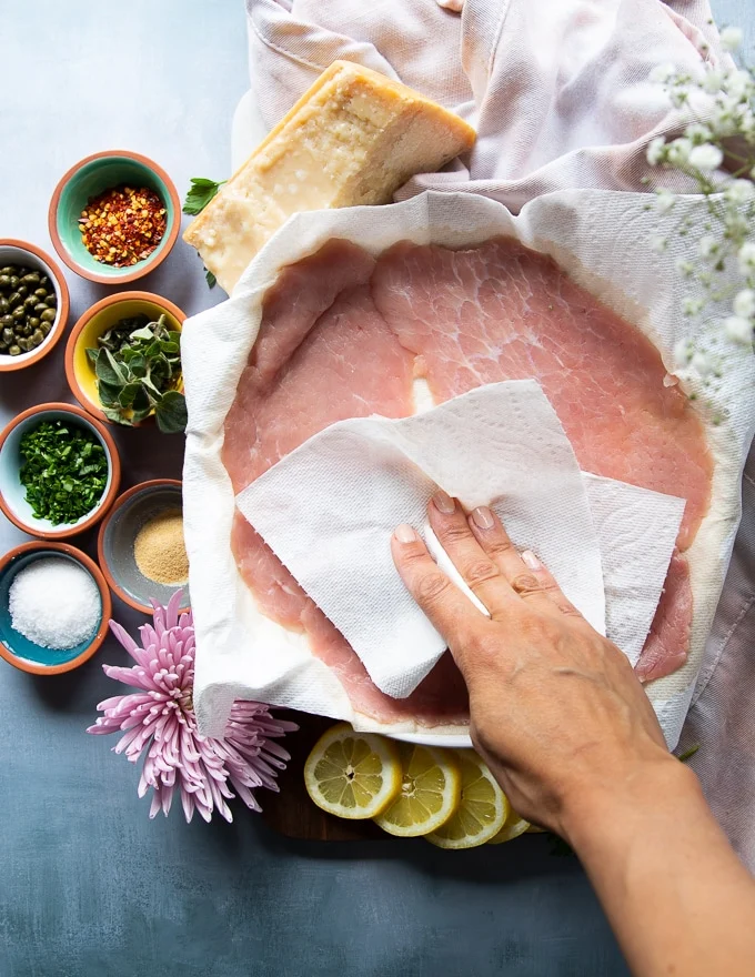 Thin slices of veal scallopini on a plate and a hand patting dry the veal using a paper towel