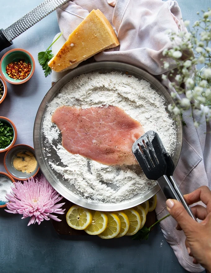 A hand holding a slice of veal scallopini using a tong and dredging it in the flour bowl