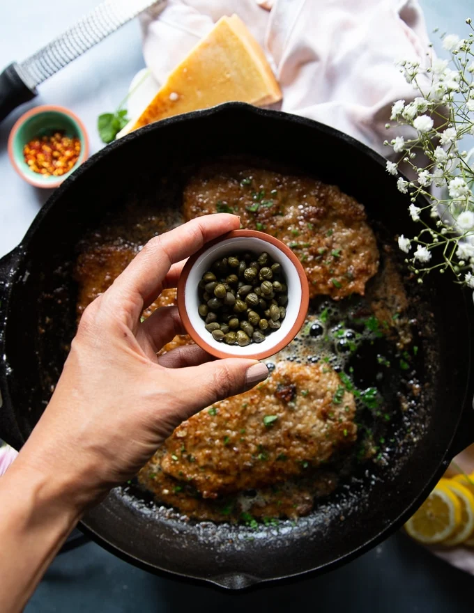 A hand adding in some capers to the piccata sauce