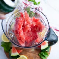 A hand pouring some soda drink into the blender with the frozen watermelon to blend into a slushie drink