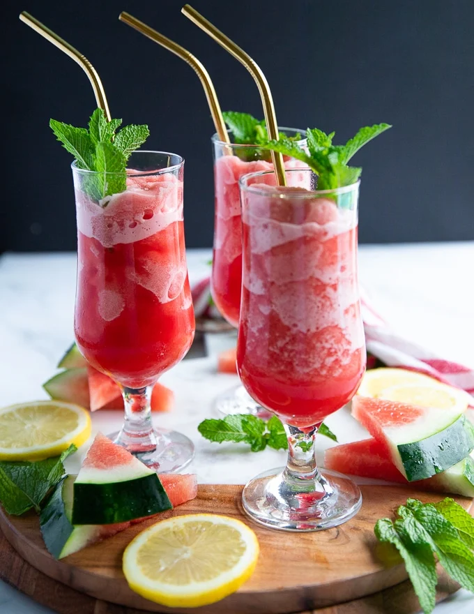 three cups of watermelon slushie with straws and mint sprigs on a board surrounded by lemon slices