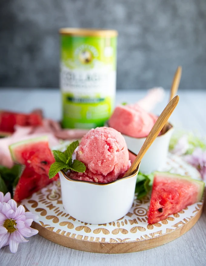 close up of one cup of watermelon sorbet with two scoops in a white bowl and a wooden spoon, and a garnish of mint