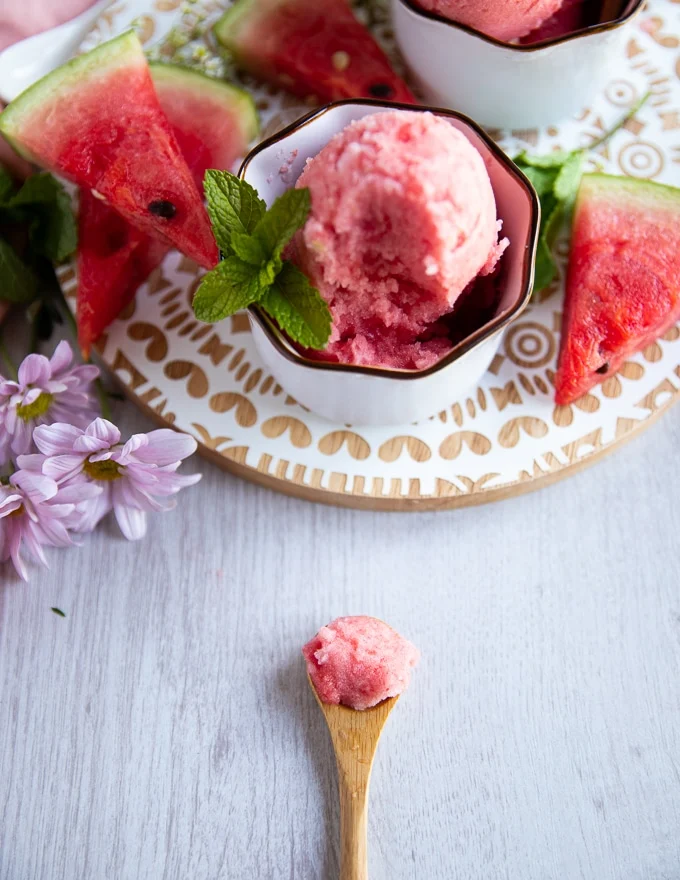 a wooden spoon with some watermelon sorbet on the board and a bowl of sorbet at the back
