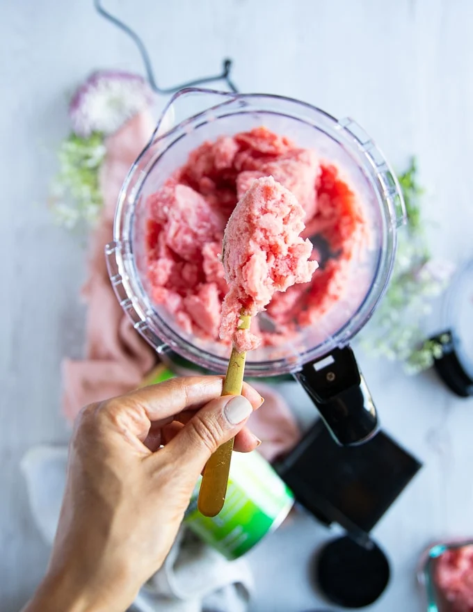 a hand holding off a spoon showing the processed watermelon sorbet recipe over the bowl of food processor