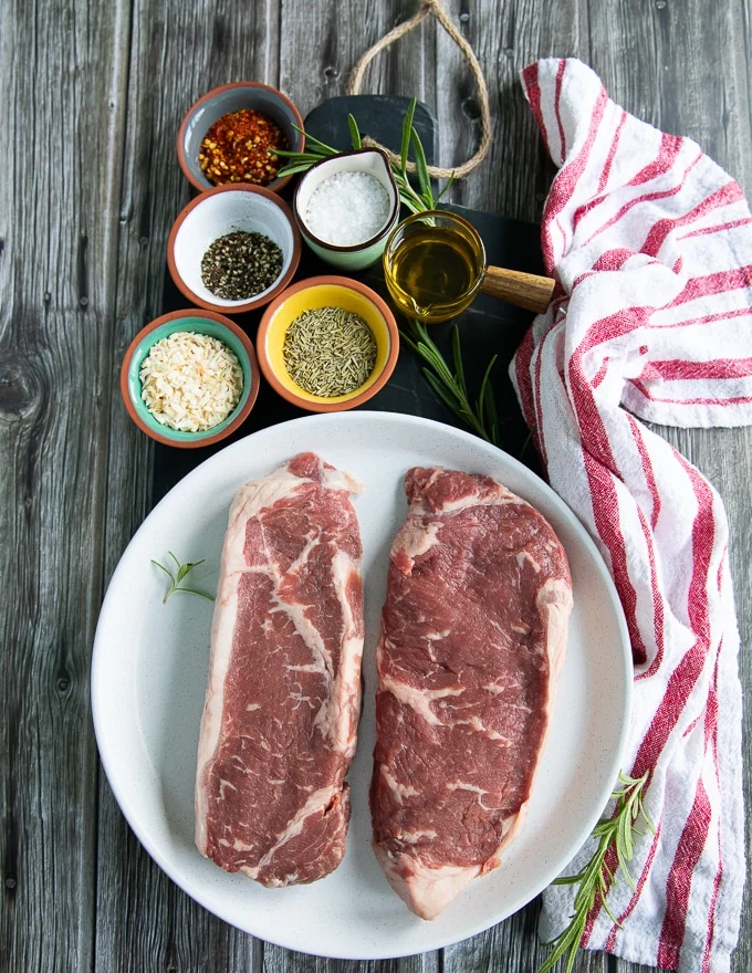 ingredients to cooking NY strip including two pieces of NY strip steak, seasoning on the side and olive oil.