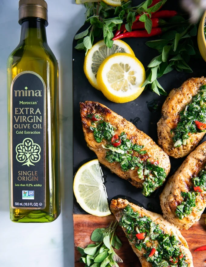 A black board with chicken chimuchurri surrounded by lemon slices, fresh oregano and a bottle of olive oil