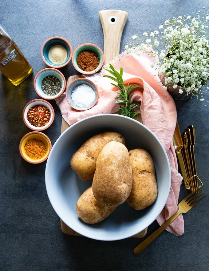 Ingredients for the air fryer baked potatoes including russet potatoes in a bowl. seasonings in small bowls and olive oil