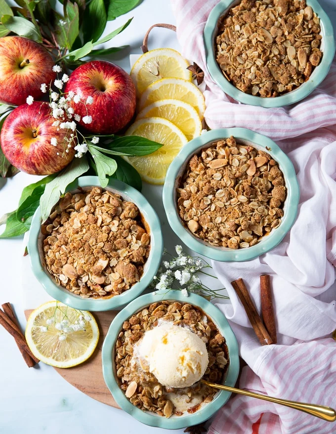 A set of four baked apple crumbles on a board surrounded by a spoon and vanilla ice cream on one of them, some apples, lemon slices and cinnamon sticks