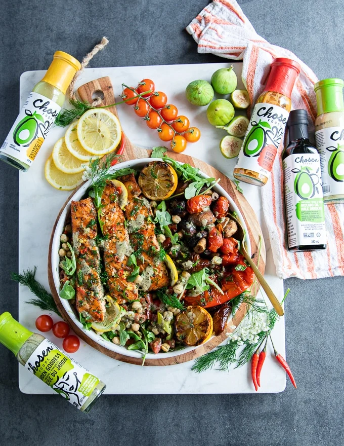 grilled salmon served on a plate surrounded by chickpea salad, some lemon slices, grilled veggies and a collection of dressings