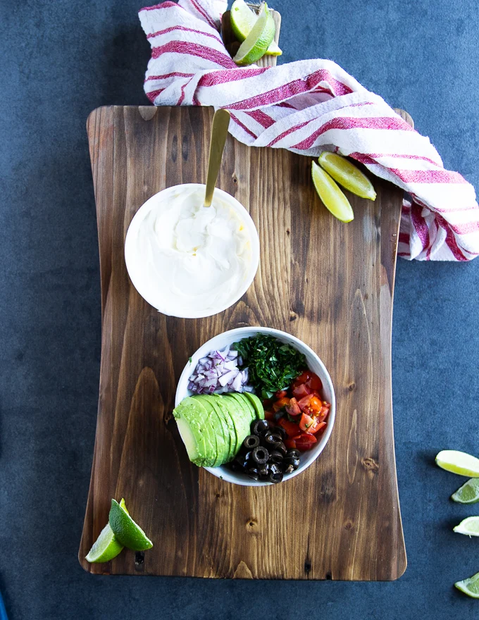 A colecyion of different nachos topping to sprinkle of the baked jalapenos including black olives, tomatoes, cilantro, onions, avocado and sour cream