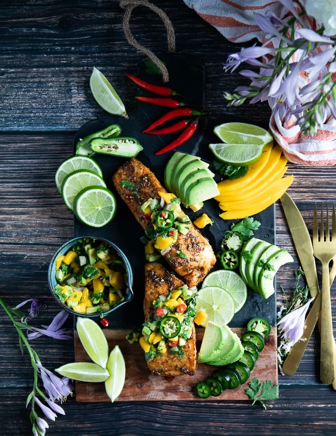 two pieces of air fryer salmon on a board surrounded by lime wedges, avocado slices, mango, avocado salsa,