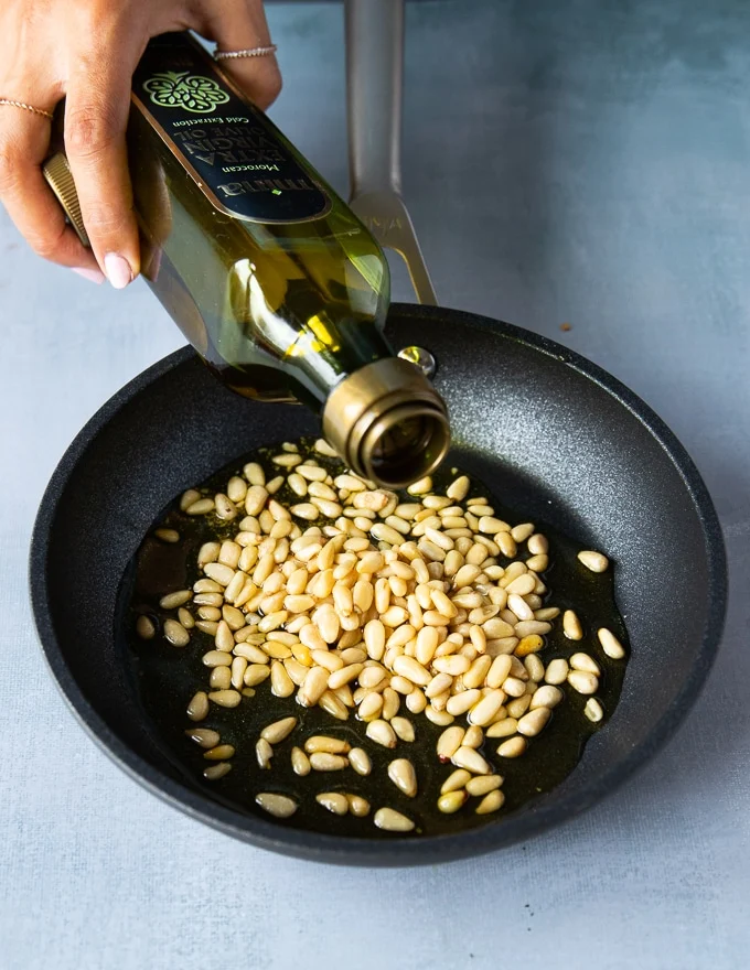 Raw Pine nuts in a skillet and a hand pouring over some olive oil over the nuts
