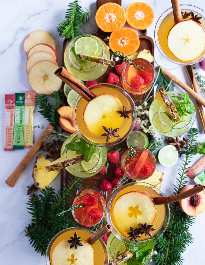 a large tray with a collection of al the mocktail recipes and different garnishes such as fresh raspberries, apples, pineapple slices, fresh mint, lime, fresh thyme and whole spices