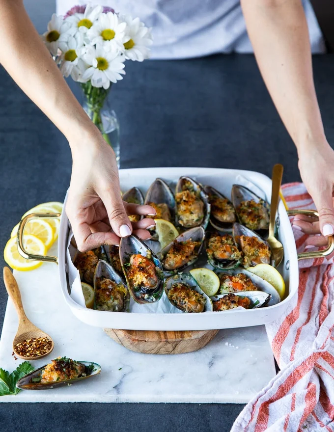 A hand holding a green mussel over a tray of cooked new zealand mussels to show the size and texture