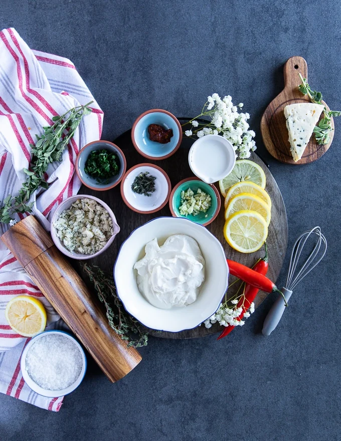 All blue cheese ingredients on a wooden board surrounded by a tea towel. A bowl of sour cream, a wedge of blue cheese, lemon, garlic, fresh herbs, salt and pepper and milk