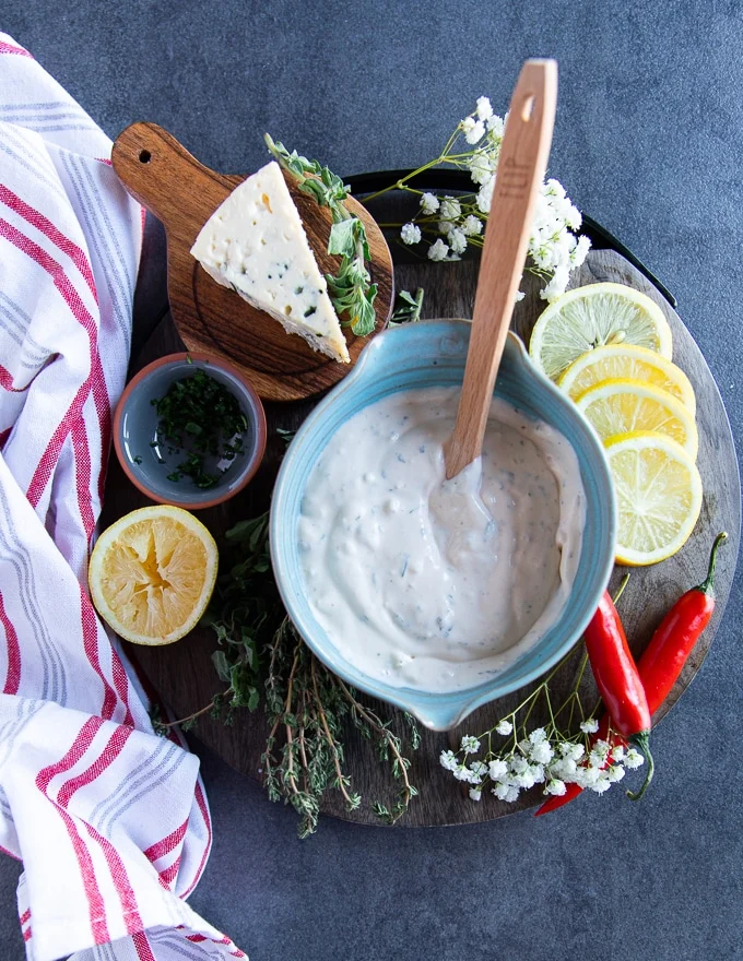 A bowl of blue cheese dressing surrounded by lemon slices, a blue cheese wedge and some fresh herbs over a wooden board and a red tea towel