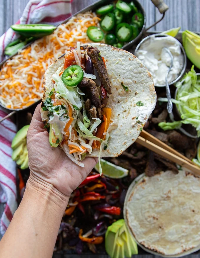 a hand holding an assembled fajita with tortilla, cooked veggies and meat, some jalapenos, cheese, salsa, avocado, sour cream