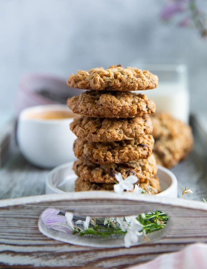 arranged and stacked oatmeal cookies on a white plate surrounded by a cup of milk and a cup of coffee and more chocolate chunks