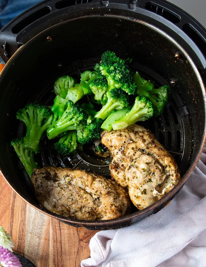 chicken breasts in an air fryer basket fliped half way and ready to continue cooking. A hand adding in some broccoli next to the chicken breasts to air fry them with the chicken to make a full meal