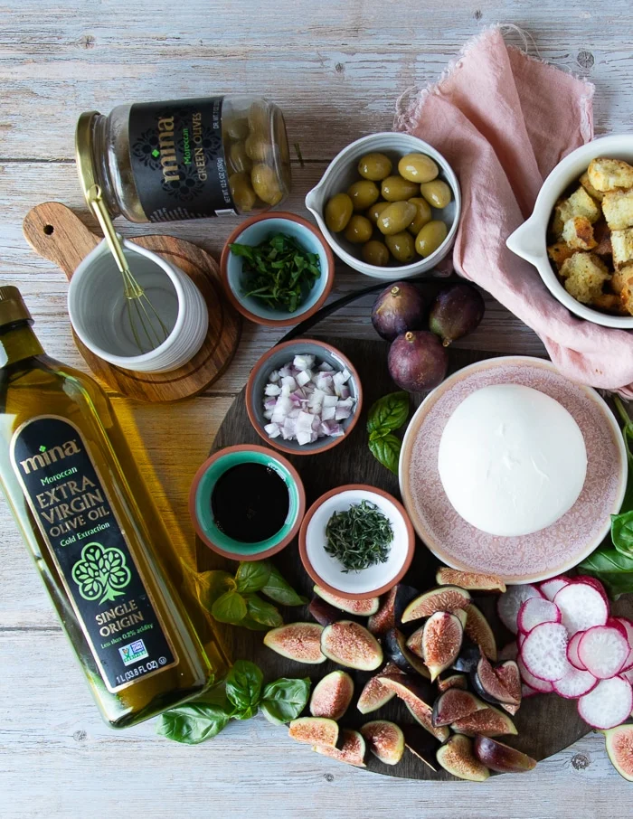 burrata salad ingredients including a plate of burrata cheese, some fresh thyme, and basil. diced shallots, green olives, arugula, fresh figs, radishes, a bowl of croutons