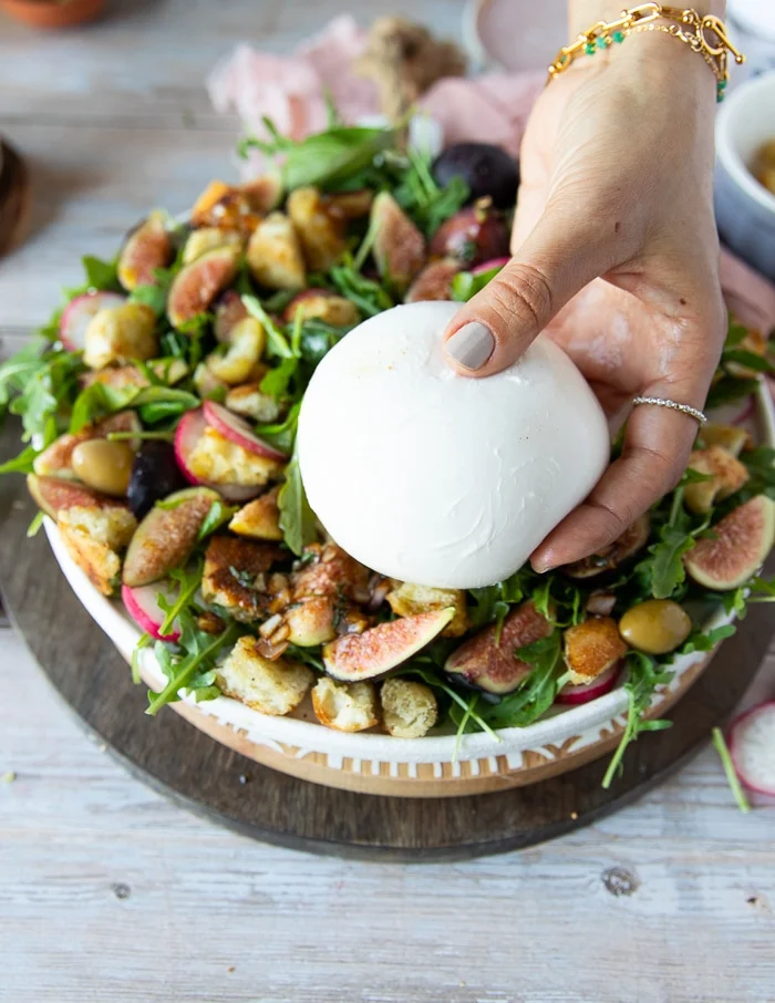 a hand holding a ball of burrata cheese ready to place it overt the salad