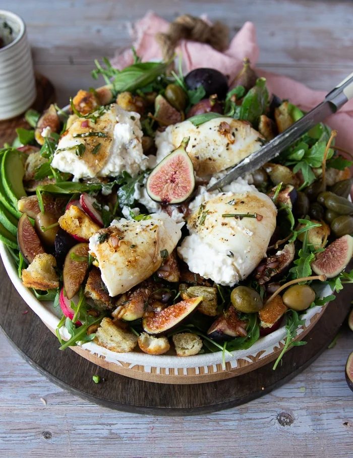 A hand holding a knife and cutting the burrata cheese on the salad