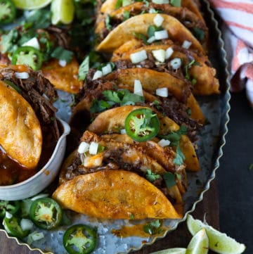 A row of lamb birria tacos arranged on a metal plate sprinkled with cilantro, onions and jalapeรฑos