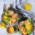 two sushi bowls on a board with wooden sticks. The bowls each have spicy salmon, sushi rice, edamame, avocado, cucumber, mango, pikcled gnger and a drizzle of spicy mayo