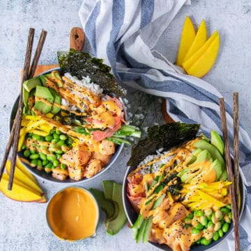 two sushi bowls on a board with wooden sticks. The bowls each have spicy salmon, sushi rice, edamame, avocado, cucumber, mango, pikcled gnger and a drizzle of spicy mayo