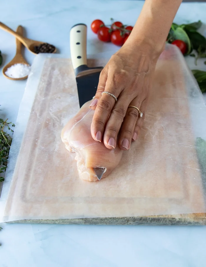 a hand using a sharp knife to cut through the chicken breast and butterfly it