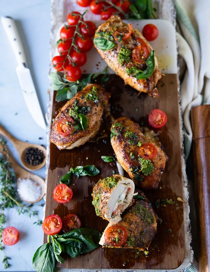 A large wooden board with caprese chicken topped with pesto, tomatoes and stuffed with tomatoes, mozzarella and basil leaves. Once piece of chicken cut in half to show the inside