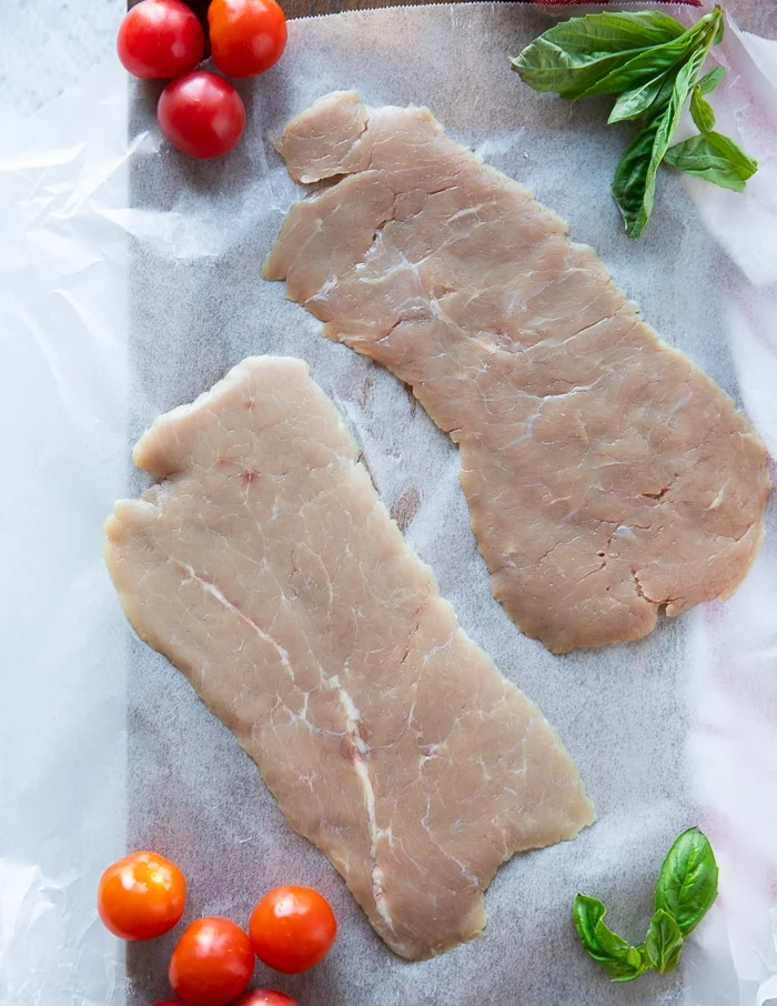 thinly sliced milk fed veal cutlets on a white paper surrounded by fresh basil leaves and fresh tomatoes