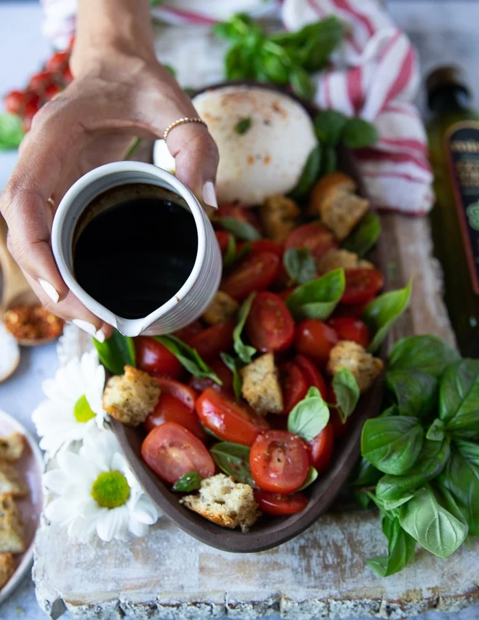 a hand holding a small pouring jug of balsamic syrup mixed with olive oil and about to pour it over the burrata caprese